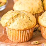 A close-up of freshly baked pistachio muffins on a wooden surface with a bowl of pistachios in the background, showcasing the deliciously textured tops and vibrant green hues.