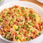 A bowl of spam fried rice mixed with vegetables, diced ham, and scrambled eggs, with a fork resting on the side.