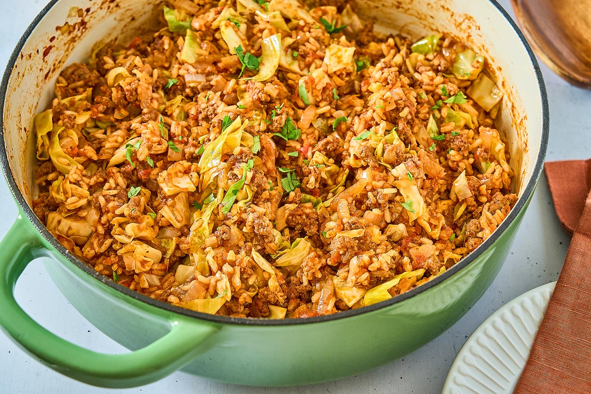 A green cast iron pot filled with a cooked dish of ground meat, cabbage, rice, and tomatoes, garnished with chopped herbs. The pot is placed on a white table next to a white plate and a brown cloth napkin.
