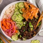 An Easy Beef Bulgogi Bowl recipe containing rice, pickled vegetables, sliced cucumbers, ground beef, and kimchi served with chopsticks on a gray surface with scattered ingredients. These Korean-inspired bowls make for a delicious and convenient meal.