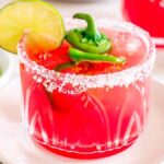 A Watermelon Jalapeño Margarita cocktail in a glass with a salt rim, garnished with a lime slice and jalapeño slices, sits on a white plate.