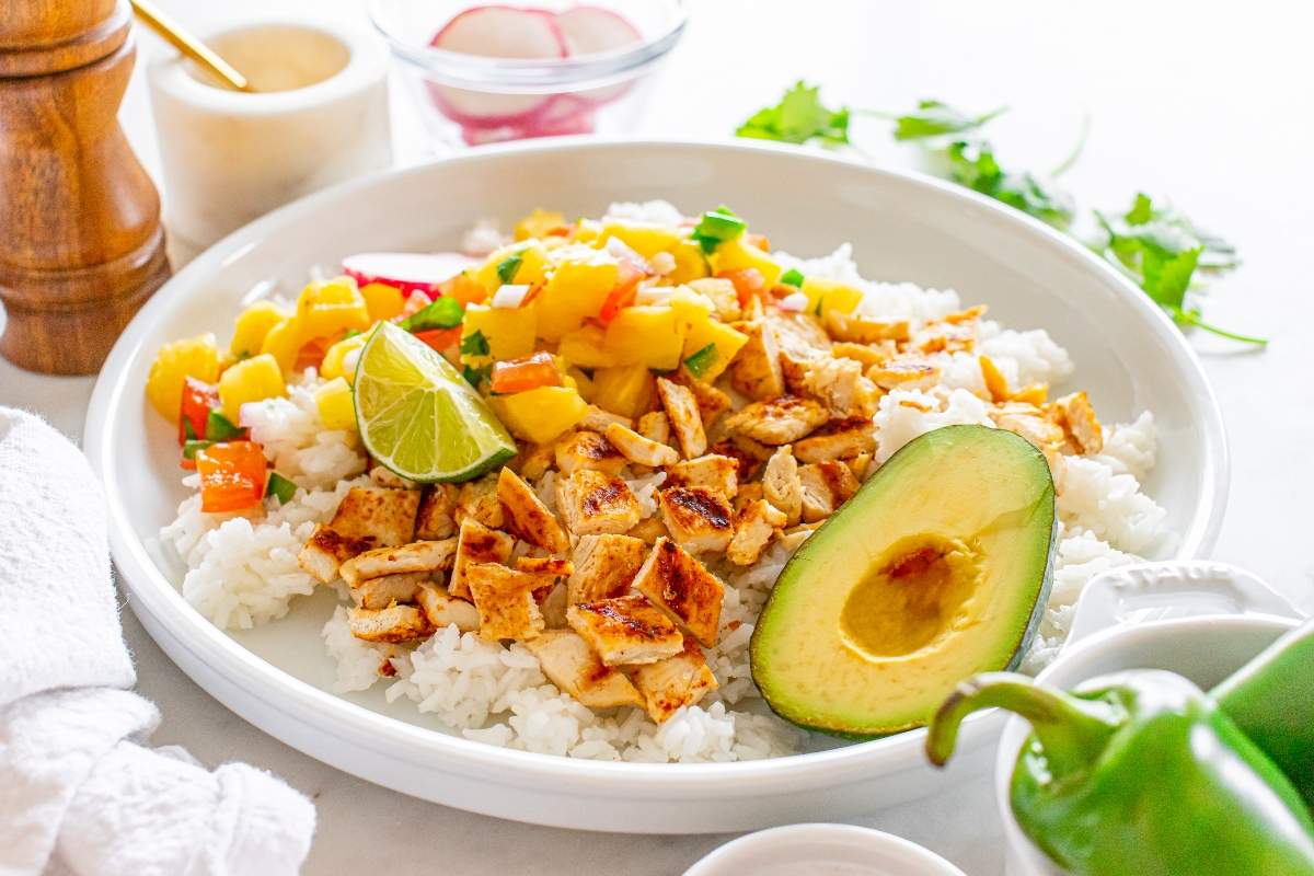 A white plate featuring grilled chicken cubes on a bed of white rice, topped with a colorful mango salsa. To the side is a halved avocado and a lime wedge. In the background, there's a glass bowl with radishes, a wooden pepper grinder, and a sprig of cilantro.