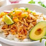 A bowl of grilled chicken with pineapple salsa on rice. Avocado halves, lime wedges, and a garnished plate in the background.