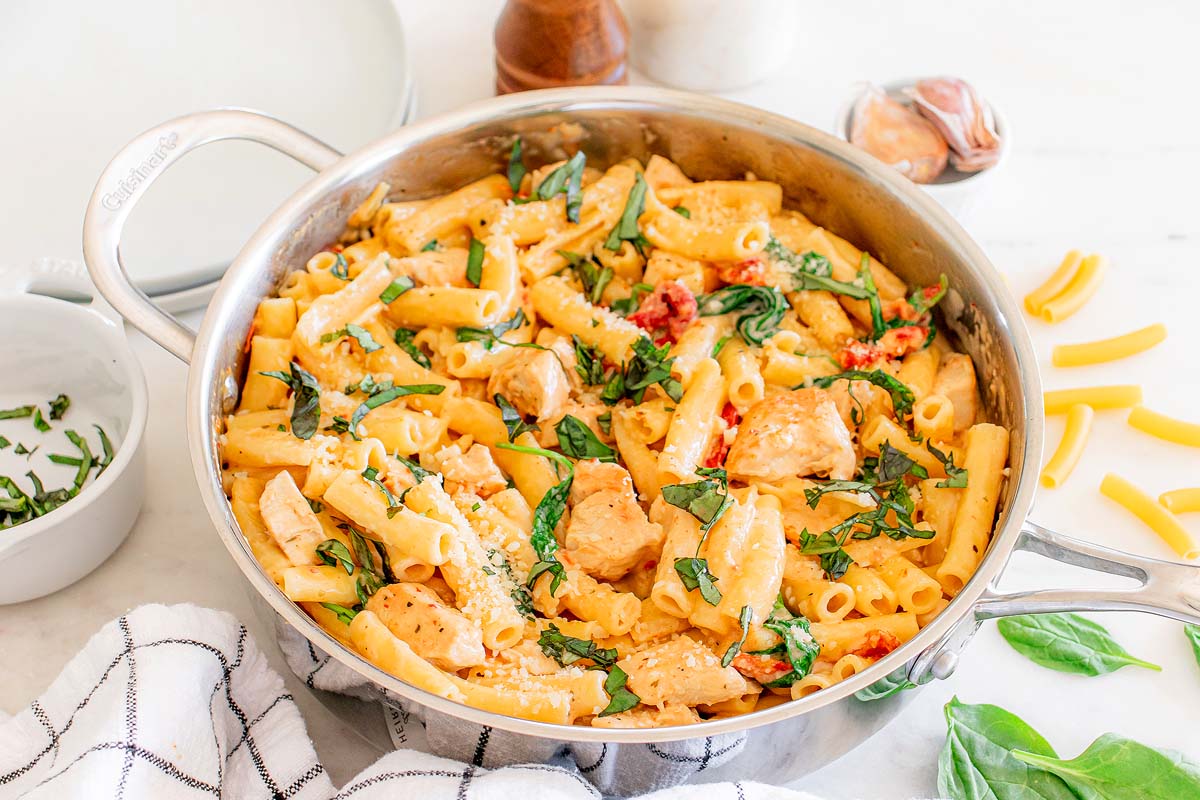 A skillet filled with creamy chicken pasta, garnished with fresh spinach and sun-dried tomatoes. Rigatoni noodles, tender chicken pieces, and a creamy sauce are visible. Fresh basil and cheese are sprinkled on top. A white dish towel and pasta pieces are nearby.