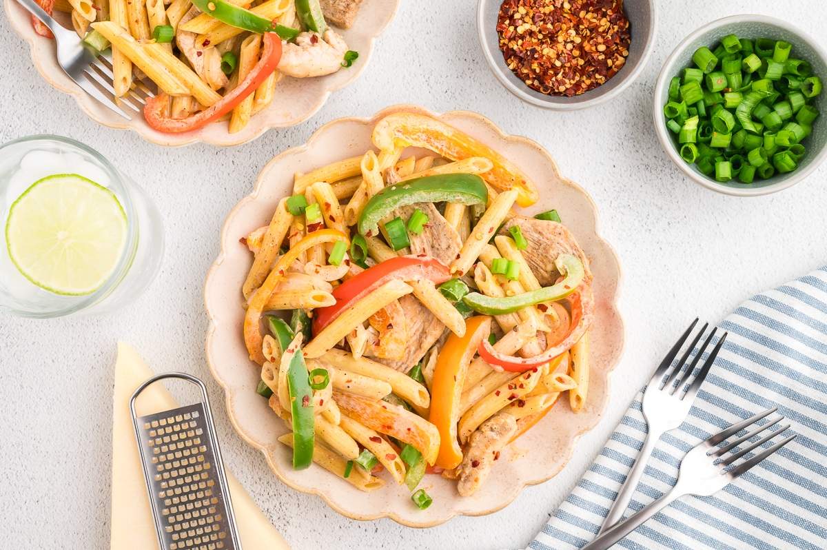 A plate of penne pasta mixed with sliced bell peppers and pieces of meat, garnished with chopped green onions, served on a scalloped-edge plate. Surrounding the plate are a glass of water with lime, small bowls of green onions and red pepper flakes, and utensils.