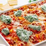 A layerless Giada sheet pan lasagna brimming with pasta and a spinach ricotta mixture sits on a marble countertop. Nearby, you'll find a pepper mill, uncooked lasagna sheets, parsley, and a bowl of grated cheese.