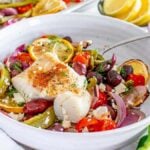 A bowl of baked sheet pan Mediterranean cod with sliced lemon, cherry tomatoes, bell peppers, Kalamata olives, feta cheese, and herbs is served with a fork on a white table.