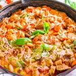Italian cannellini bean and sausage skillet dinner topped with grated cheese and basil leaves. Fresh cherry tomatoes, basil, and a striped cloth are placed around the skillet.