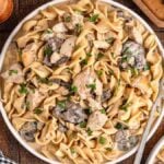 A plate of creamy chicken stroganoff, with mushroom gravy and a parsley garnish, placed on a wooden table with a fork, checkered cloth, and additional ingredients nearby.