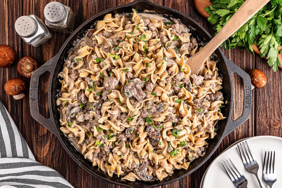A cast-iron skillet filled with creamy beef stroganoff pasta garnished with chopped herbs. A wooden spoon rests in the skillet. The table is set with a striped cloth, salt and pepper shakers, mushrooms, parsley, plates, and forks.