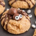 Halloween Peanut Butter Spider Cookies decorated with chocolate drizzle, edible eyes, and chocolate balls resembling spiders. The cookies are arranged on a dark surface.