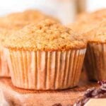 Close-up of several oatmeal raisin muffins arranged on a wooden surface, with oats, raisins, and a hint of cinnamon in the foreground.
