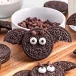 Cookies decorated to look like bats with eyes made from candies and ears made from slivers of additional cookies sit on a wooden surface. Bat Oreos, other unadorned cookies, and a glass of milk are also visible.