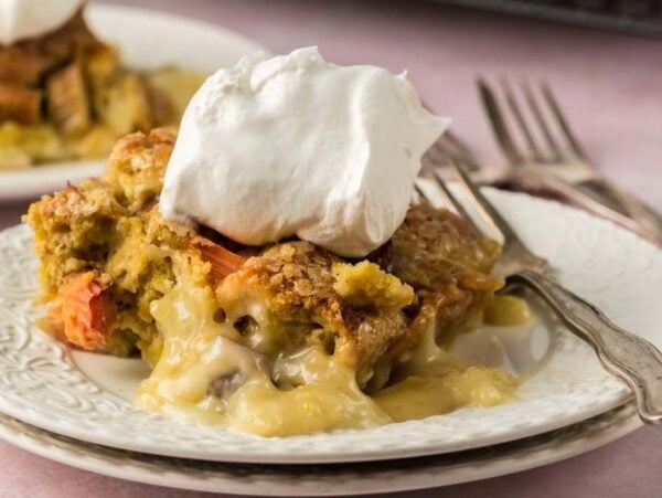 A slice of rhubarb custard cake topped with whipped cream on a decorative white plate, with another slice in the background, accompanied by a fork and knife.