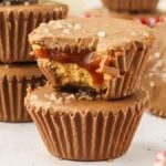 Stack of Salted Caramel Peanut Butter Cups, one with a bite taken, revealing gooey caramel and crumbly layers. Topped with coarse salt flakes, against a background with more candy and a checkered cloth.