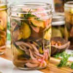 Jars of spicy dill pickles, including sliced cucumbers and onions, are arranged on a wooden surface with fresh dill.