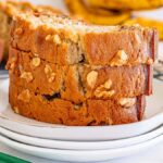 Three slices of Starbucks copycat banana bread stacked on a white plate with a cooling rack and bananas in the background.