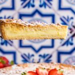 A hand holds a slice of torta della nonna over a whole cake topped with strawberry slices. Blue and white patterned tiles are in the background.