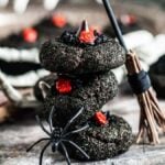 Stack of Halloween-themed witch hat cookies adorned with red candy roses, surrounded by a toy spider, a miniature broomstick, and skeletal hands.