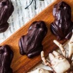 Five Harry Potter chocolate frogs and a skeletal hand decoration are placed on a wooden board and crumpled book pages, creating an enchanting scene.