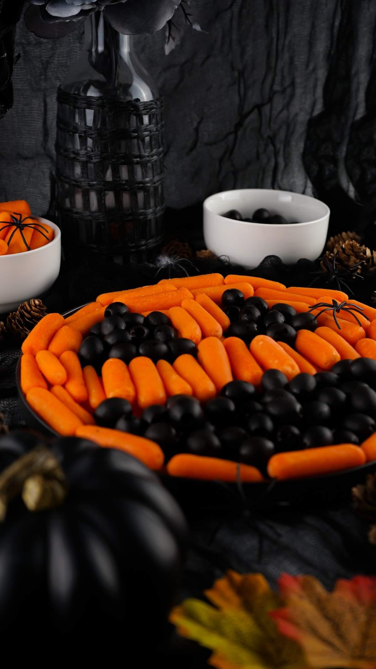 A Halloween veggie board featuring a pumpkin face made from baby carrots and black olives. The background boasts black decorative items, two small bowls, and a black pumpkin.
