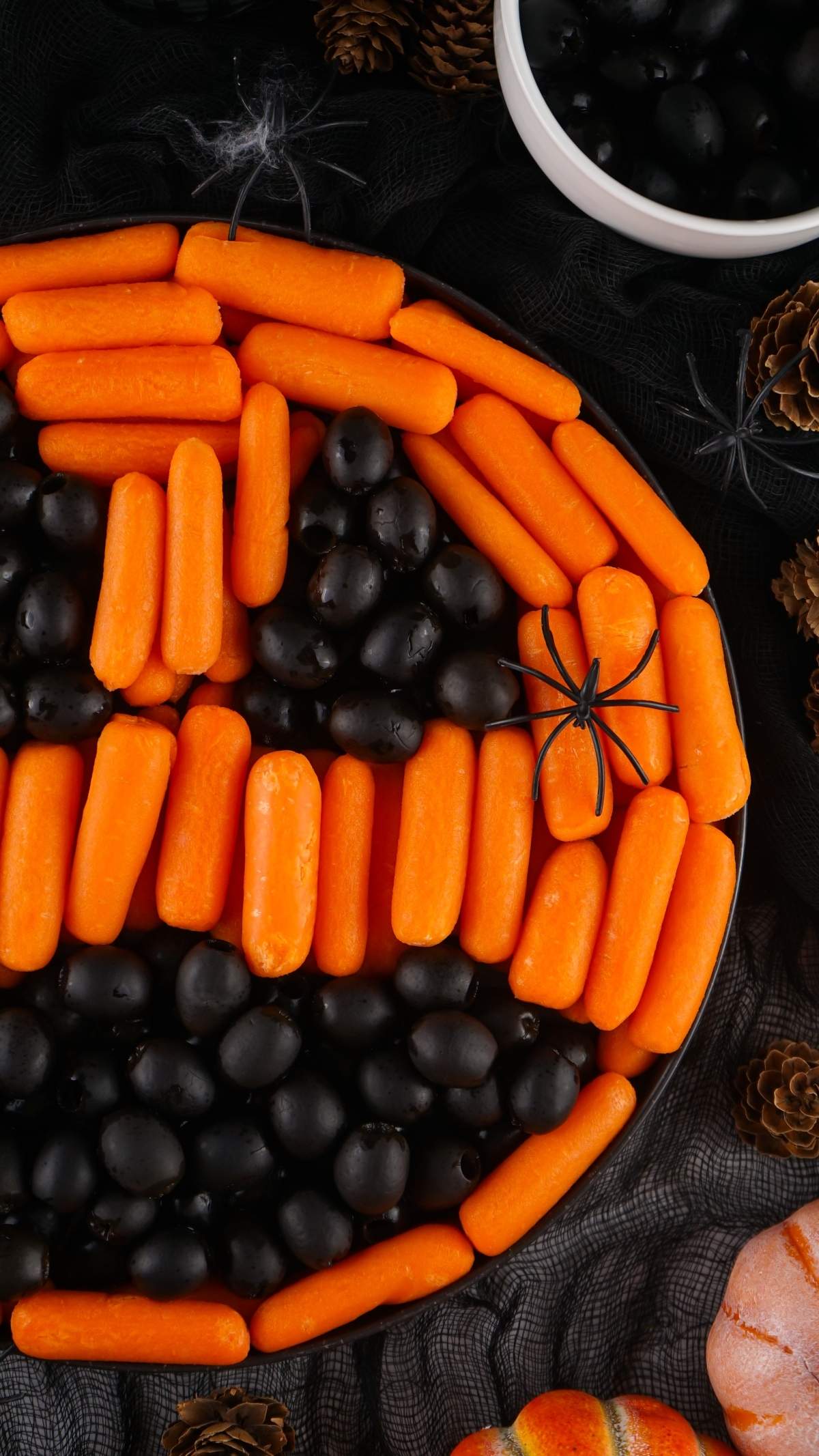 A A Halloween veggie platter featuring a tray of baby carrots and black olives arranged to create a jack-o'-lantern face, surrounded by autumn decorations and small pumpkins.with carrots and black olives arranged on a platter to look like a jack-o'-lantern face, with additional olives in a bowl nearby. A plastic spider decoration adds a spooky touch.
