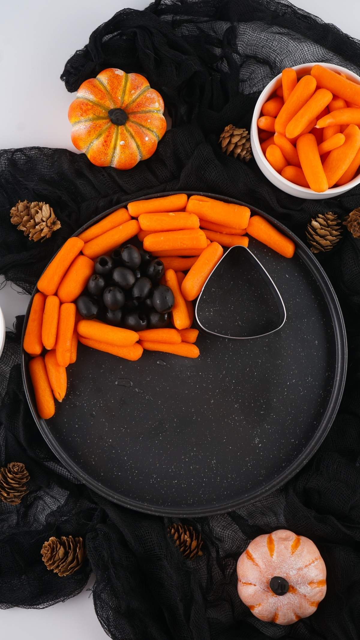 A Halloween veggie platter featuring a round black tray with baby carrots and black olives arranged in a crescent shape, surrounded by small pumpkins, pinecones, and black fabric. Two bowls of baby carrots are positioned nearby for extra nibbling.