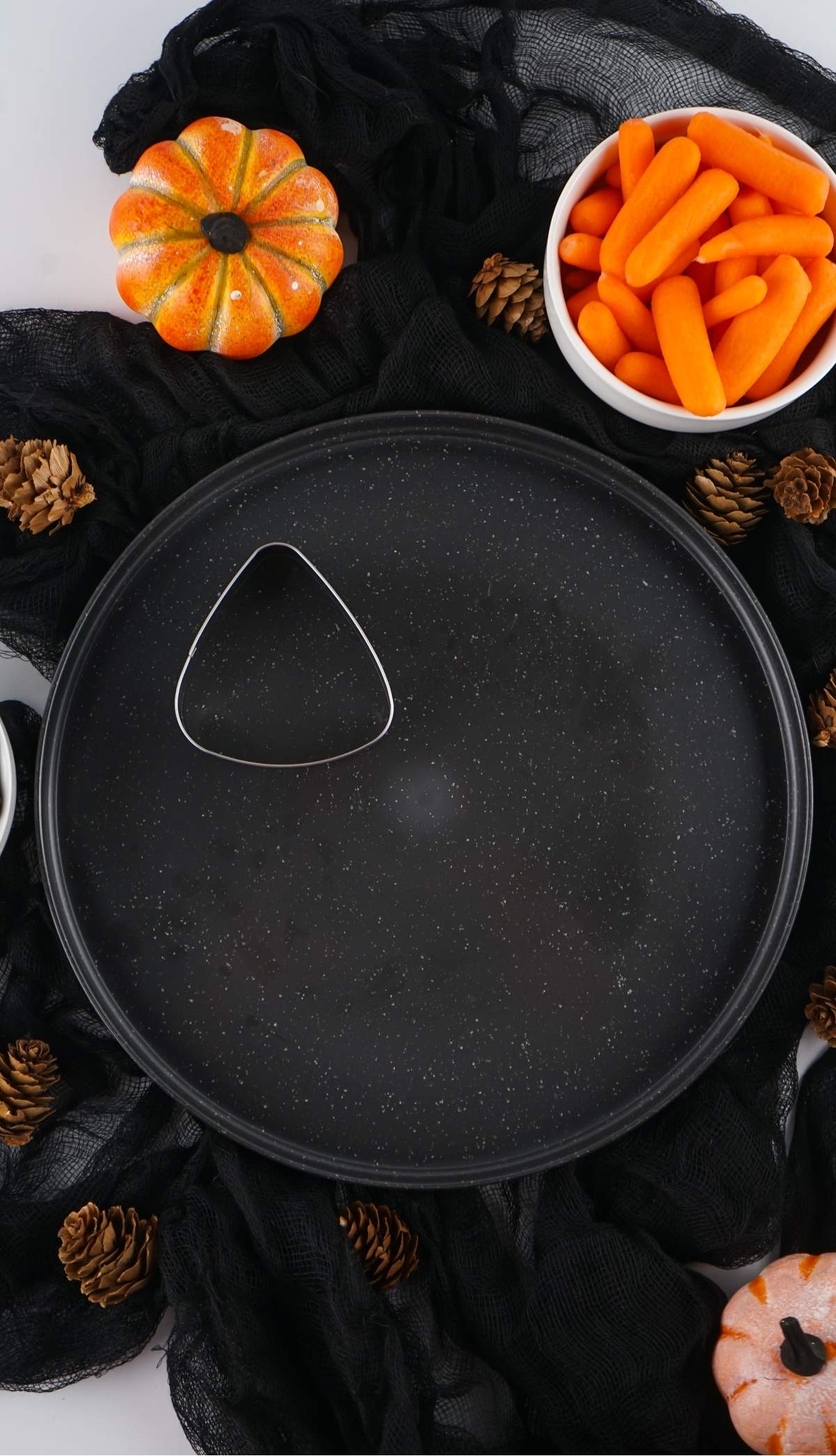 A black round tray with a triangular dip bowl is surrounded by small pumpkins, pinecones, a bowl of baby carrots, and a black cloth, creating the perfect Halloween veggie platter.