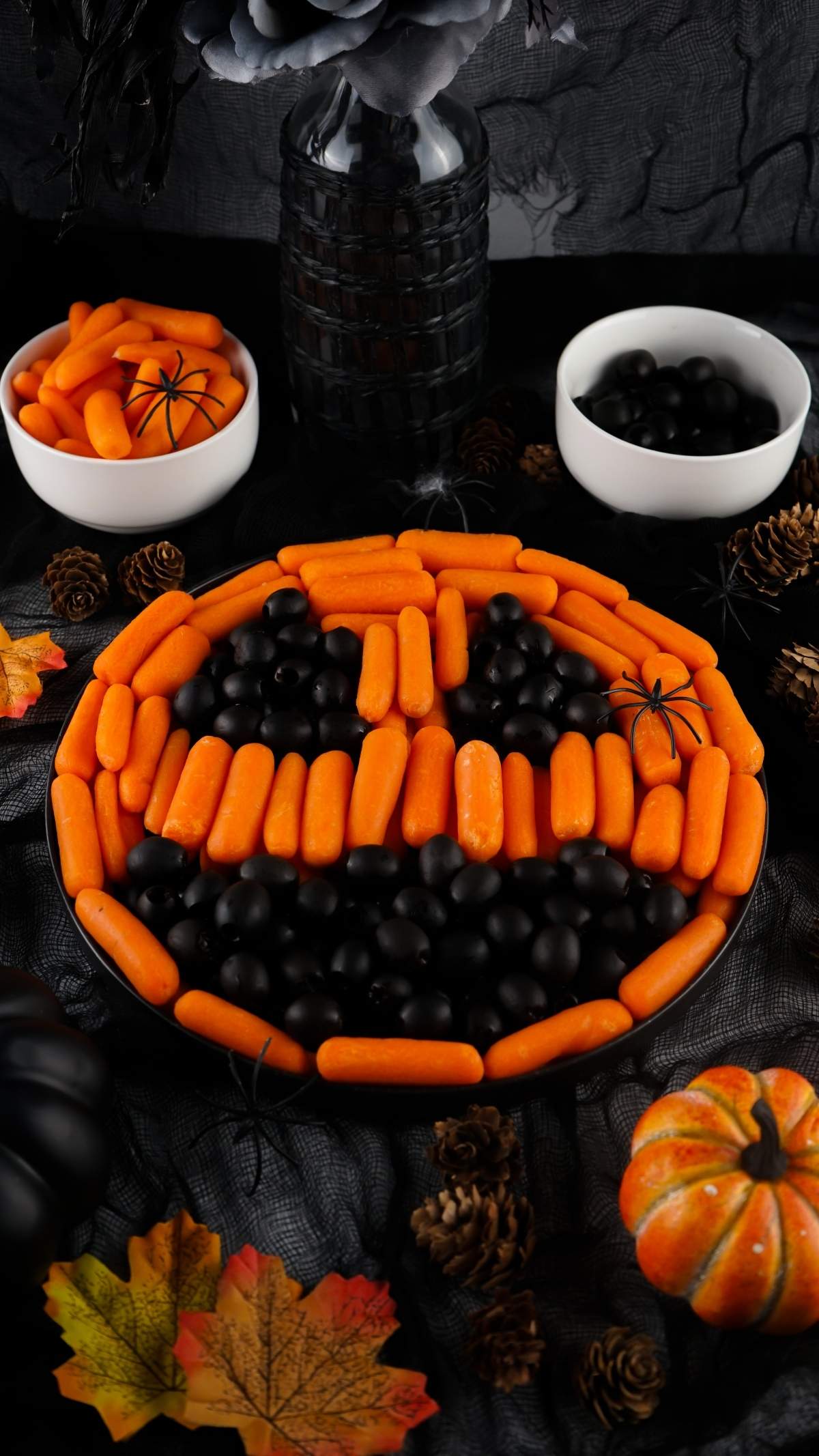 A Halloween jack-o-lantern veggie platter arranged to resemble a jack-o'-lantern face, with orange carrots forming the pumpkin and black olives as eyes, nose, and mouth. Surrounded by festive decor.