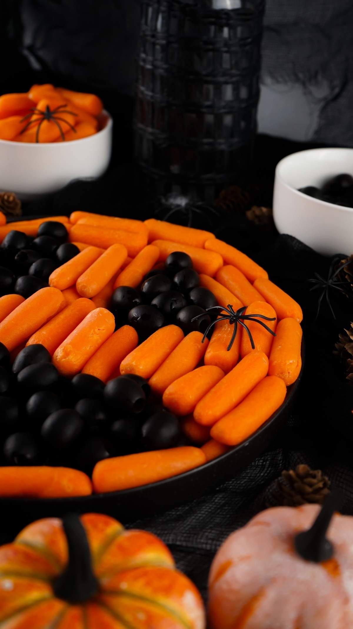 An easy Halloween veggie tray features alternating rows of baby carrots and black olives, with fake spider decorations. Adjacent bowls contain more baby carrots and black olives. Pumpkins are placed in the foreground.