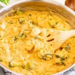 A pot of creamy cheesy chicken broccoli orzo, stirred with a wooden spoon. A pepper mill and a small bowl with a golden spoon are in the background.