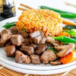 A plate of hibachi steak cubes, fried rice, and mixed sautéed vegetables, with chopsticks and a bottle of soy sauce in the background.