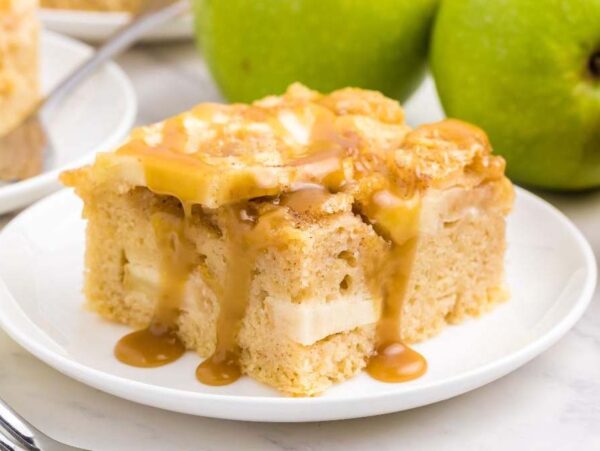 A slice of apple sheet cake with caramel sauce drizzled on top, served on a white plate with whole green apples and a glass of caramel sauce in the background.