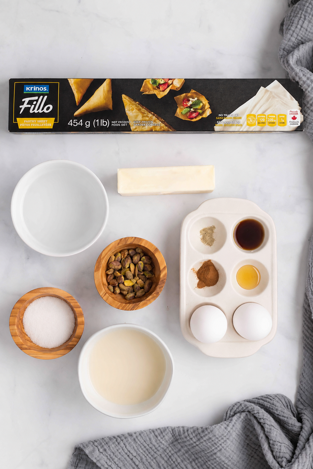 Fillo dough, block of butter, eggs, spices, milk, sugar, pistachios, and two bowls on a marble surface.
