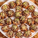 A plate of caramel apple pretzel bites is topped with chocolate drizzle and sprinkled with chopped nuts and candy pieces. Additional caramel candies and pretzels are scattered around the wooden surface.