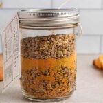A glass jar filled with layered lentils and spices, perfect for making coconut lentil curry soup, sits on a countertop with a tag attached to its neck. A piece of flatbread, a spoon, and a white bowl are in the background.