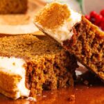 Two slices of frosted gingerbread loaf on a brown plate, with one slice leaning against the other, and red berries blurred in the background.