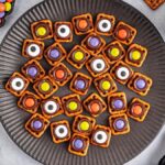 A black plate with Halloween pretzel snacks topped with colorful candies and candy eyes. Surrounding items include Halloween-themed black and white pumpkins, wrapped purple candies, and a bowl of M&M-like candies.