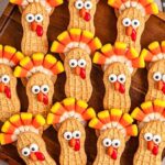 A wooden tray displays Nutter Butter turkey cookies decorated with candy corn feathers, candy eyes, and red candy beaks. Pinecones and additional cookies are visible in the background.