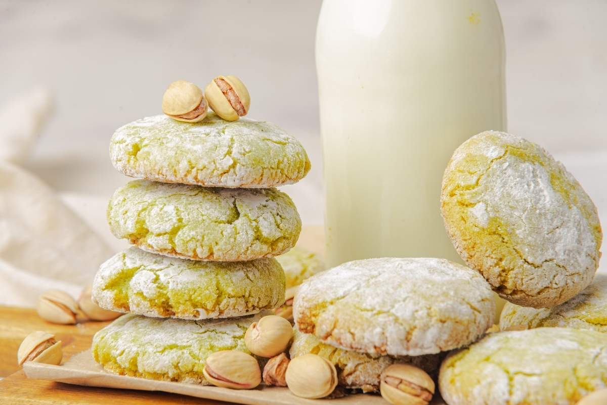 A stack of green Italian pistachio cookies and several loose cookies are displayed with scattered pistachios on a wooden surface. A bottle of milk is in the background.