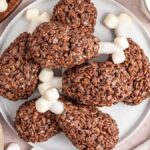 A plate of chocolate rice crispy treats shaped like drumsticks, with mini marshmallows at the ends to resemble bones. A bowl of marshmallows, a wooden tray with treats, and a glass of milk are nearby.