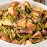 A white bowl filled with slow cooker ham, green beans, and red potatoes, with a spoon on the right side.