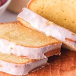 A loaf of sliced sour cream pound cake with white icing sits on a wooden cutting board next to a bowl of grapes.