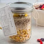 A labeled mason jar filled with dry ingredients for taco soup in a jar sits on a countertop. In the background, there is a white bowl with colorful toppings.