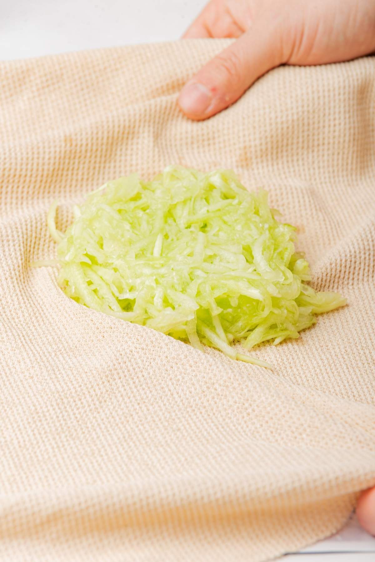 A hand holding a beige cloth with shredded zucchini, perfect for a fresh tzatziki recipe.