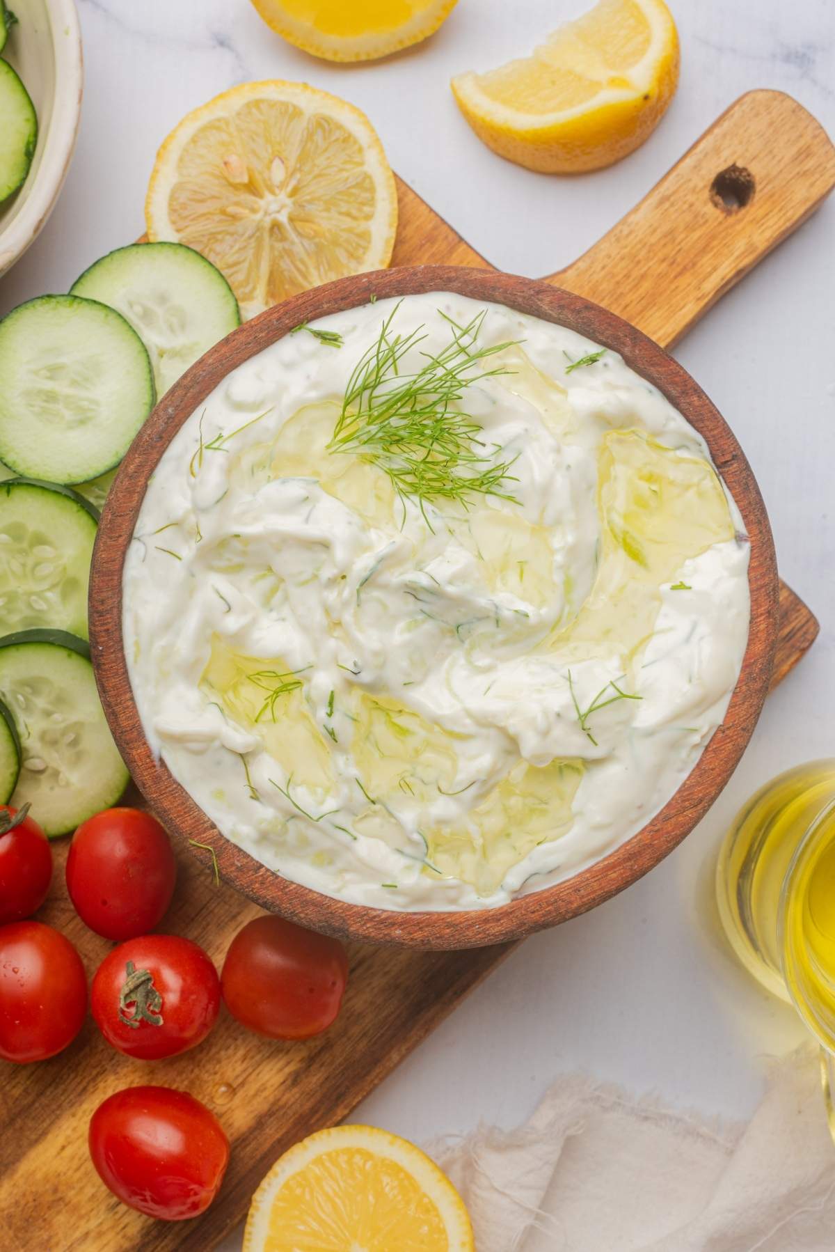 A bowl of the best tzatziki sauce is garnished with dill and surrounded by sliced cucumbers, cherry tomatoes, and lemon wedges on a wooden board.
