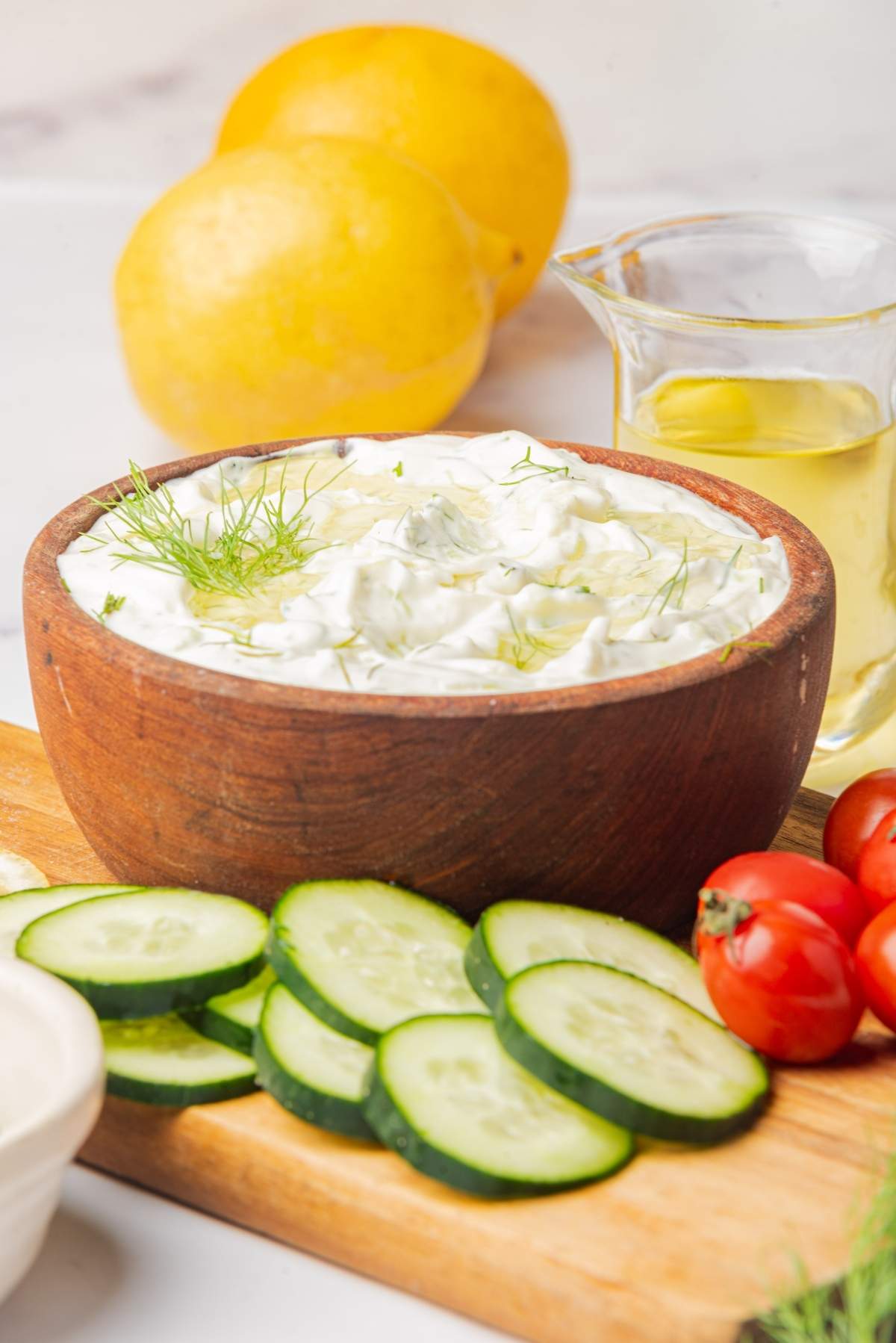 A wooden bowl filled with a classic tzatziki recipe garnished with dill sits on a cutting board with sliced cucumbers, cherry tomatoes, lemons, and a small pitcher of oil in the background.