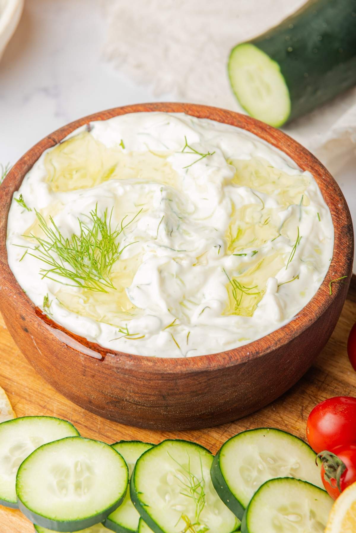 A wooden bowl filled with creamy white tzatziki sauce, garnished with dill and drizzled with olive oil. Sliced cucumbers and cherry tomatoes are placed around the bowl, making this tzatziki recipe visually appealing and delicious.