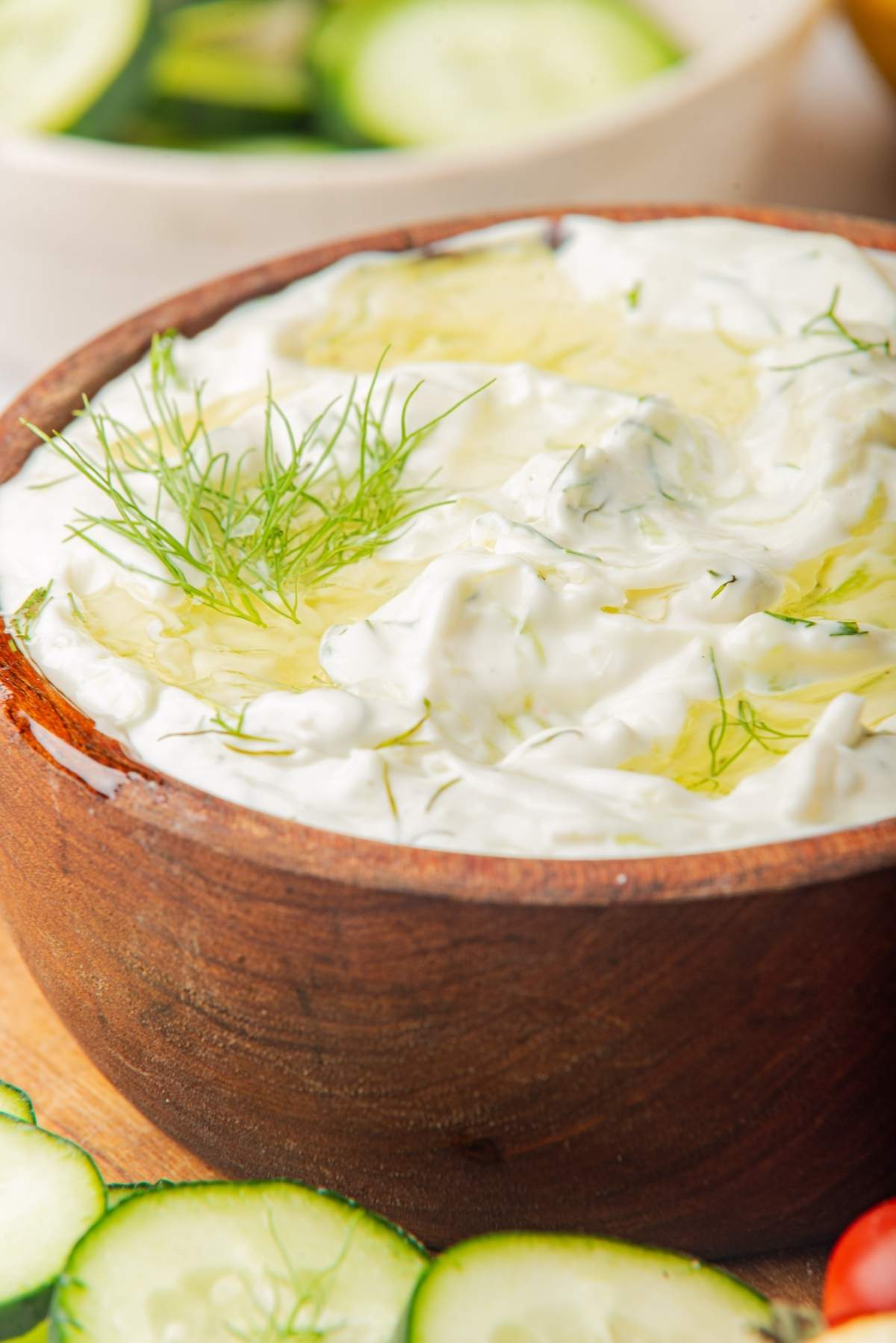 A wooden bowl filled with creamy, white tzatziki sauce garnished with fresh dill and a drizzle of olive oil. Slices of cucumber are visible in the background, perfect for trying an authentic tzatziki recipe.