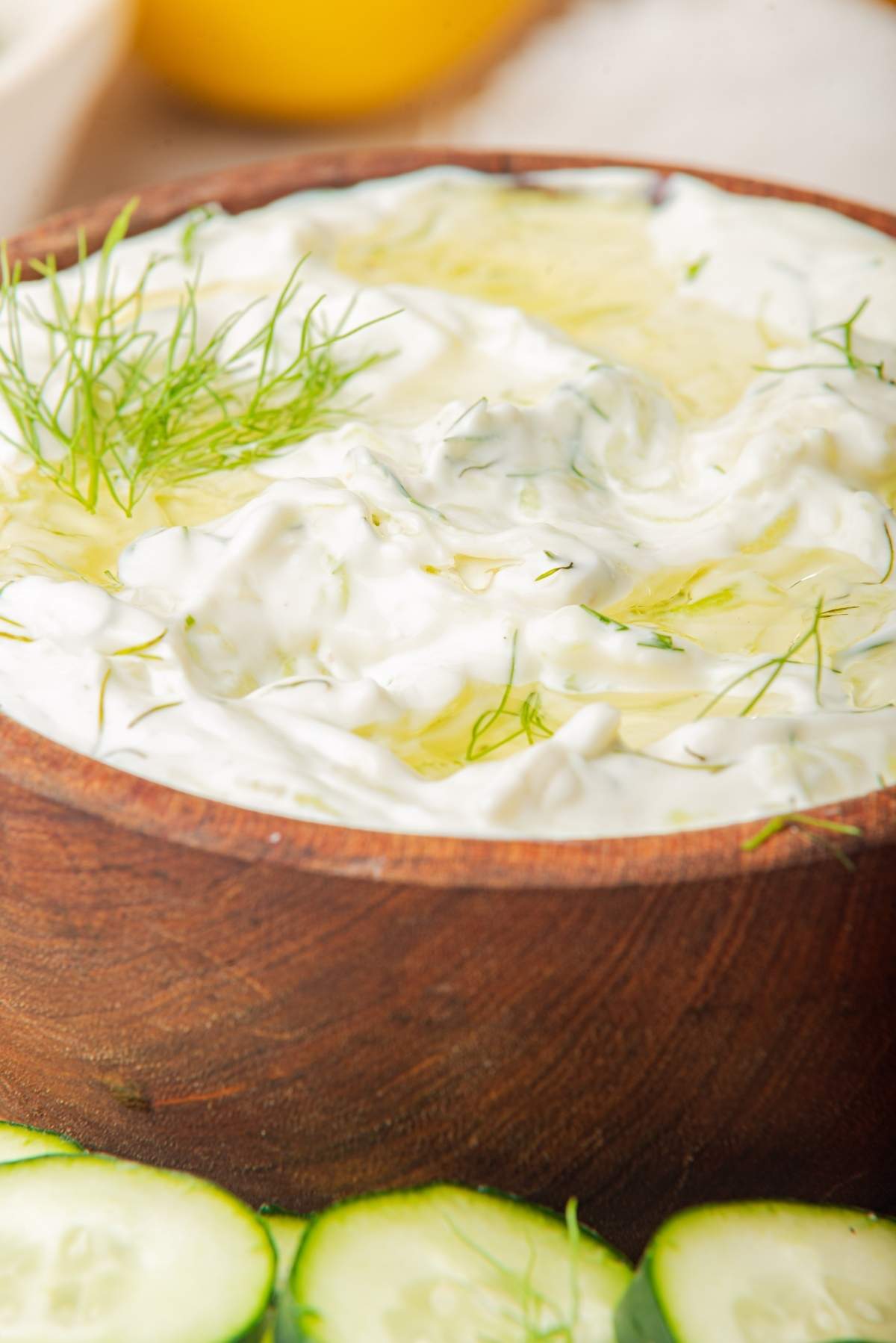 A wooden bowl filled with homemade tzatziki sauce garnished with dill, with cucumber slices in the foreground—your perfect tzatziki recipe awaits.